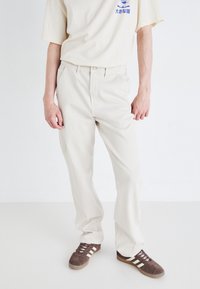 Pantaloni cargo bej din bumbac, cu croială dreapt�ă, care au buzunare frontale și fermoar, purtați cu un tricou crem și adidași maron.