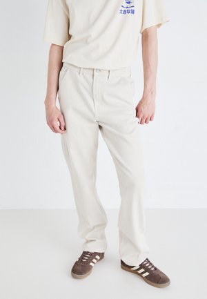 Beige cotton cargo pants with a straight leg fit, featuring front pockets and a zip fly, worn with a cream T-shirt and brown sneakers.