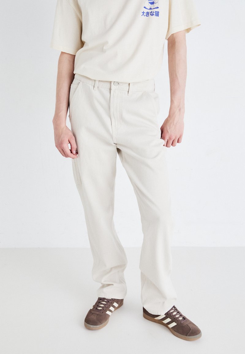 Beige bomulds cargo-bukser med lige ben, der har forlommer og lynlås, båret sammen med en cremefarvet T-shirt og brune sneakers.
