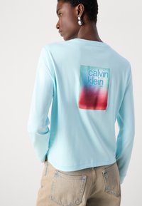 Helles blaues Langarmhemd mit einem rechteckigen Grafikdesign in Rot und Blau sowie dem Schriftzug "Calvin Klein". Dazu werden hellbeige Jeans getragen.