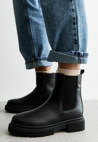 Botas Chelsea negras con paneles elásticos en los laterales, punta redondeada y suela de goma gruesa. Combinadas con jeans de mezclilla azul con dobladillo y calcetines beige.