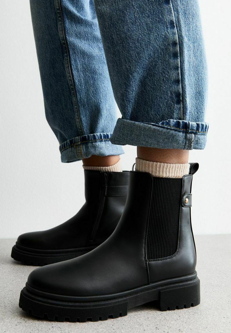 Botas Chelsea negras con paneles elásticos en los laterales, punta redondeada y suela de goma gruesa. Combinadas con jeans de mezclilla azul con dobladillo y calcetines beige.