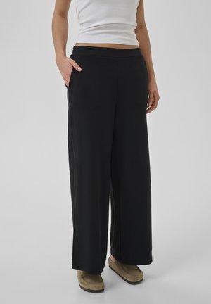 Kvinde iført sorte viddebukser, beige slip-on sko og hvid ærmeløs top, stående med den ene hånd i lommen mod en ensfarvet baggrund.