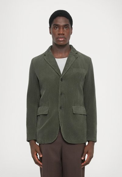 Dunkelgrüner Cordblazer mit Schalkragen, zwei vorderen Taschen, drei Knöpfen und strukturiertem Finish, getragen über einem hellgrauen Hemd.