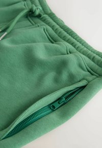 Pantalon de survêtement vert avec taille élastique, poche avant et poche zippée verte partiellement ouverte sur fond blanc.