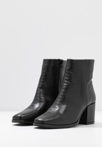 Pár černých kotníkových bot s texturovaným, hadím vzorem, bočním zipem a blokovými podpatky na bílém povrchu na šedém pozadí.