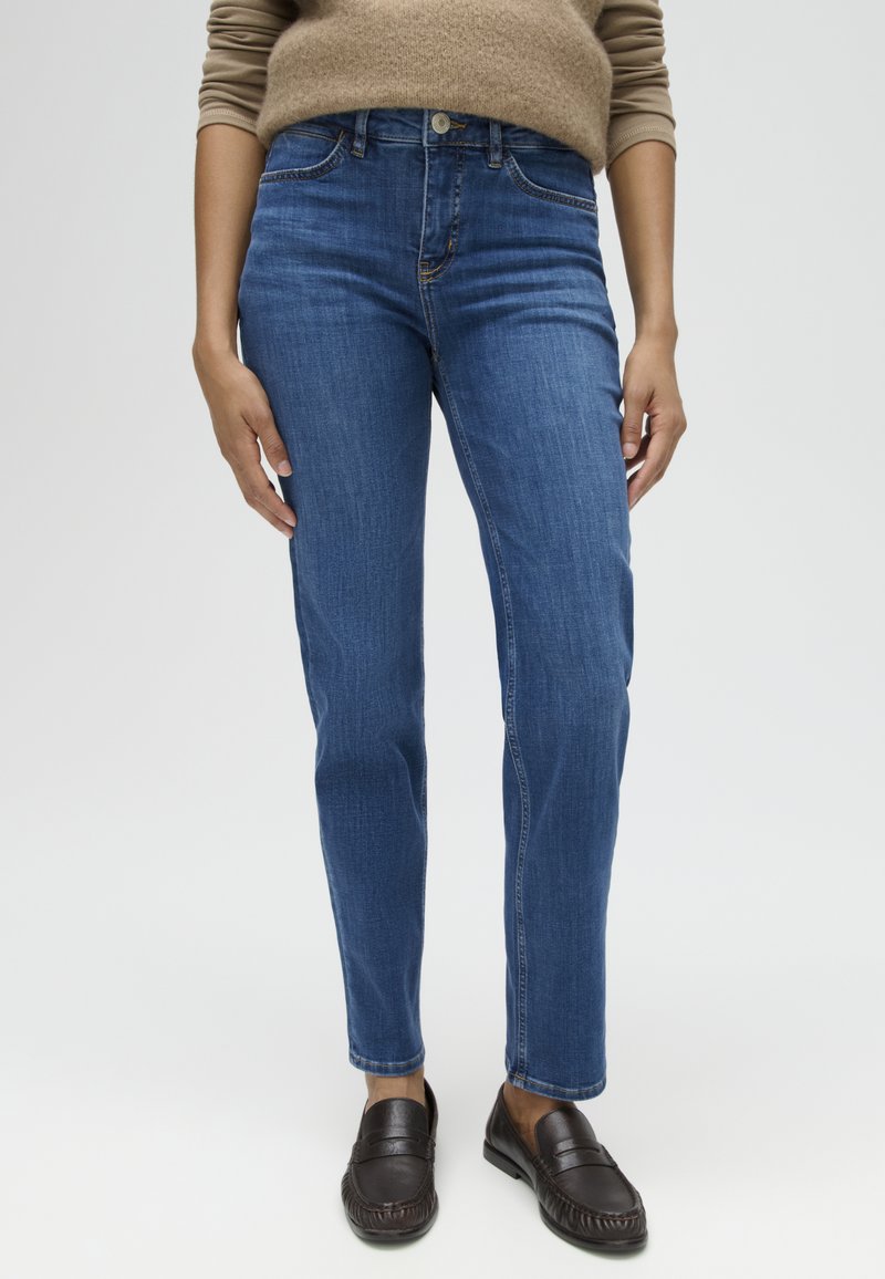 Persona che indossa jeans blu a gamba dritta, mocassini marroni e una maglia beige a maniche lunghe, in piedi contro uno sfondo neutro.