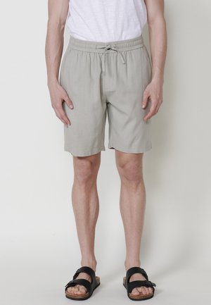 Pantalones cortos de color gris claro, hechos de una tela suave y texturizada. Presentan una cinturilla elástica con cordón y bolsillos laterales. Se llevan con sandalias negras.