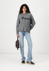 Femeie cu păr creț poartă un pulover gri cu inscripția "Paris", blugi de culoare albastru deschis, adidași negri și albi, ținând o geantă de mână maro închis pe fundal alb.