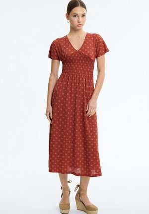 Mujer con vestido midi de manga corta de color óxido con pequeños patrones de sol y sandalias de cuña beige con tiras en el tobillo.