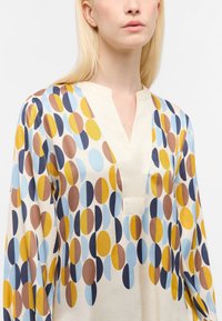 Donna che indossa una blusa con scollo a V e un motivo colorato di cerchi sovrapposti, metà e pieni, in tonalità di giallo, blu, marrone e blu navy.