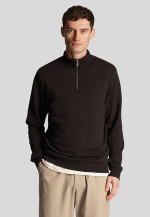 Zwarte rits-pullover gemaakt van gestructureerde stof, met lange mouwen en een klein logo op de borst, gedragen over een wit shirt.