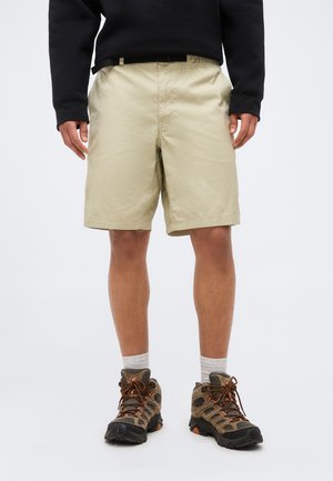 Vyras dėvi smėlinio spalvos šortus, baltas kojines, žygių batus ir juodą džemperį, stovintis ant šviesiai pilkos spalvos fono.