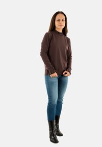 Maglietta marrone a maniche lunghe con scollatura arricciata e texture a pois, abbinata a jeans skinny blu e stivaletti neri alla caviglia.