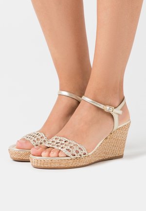 Des sandales compensées en or tressées présentent un dessus en tresse, une sangle autour de la cheville avec une boucle et une texture naturelle tissée sur la semelle compensée.