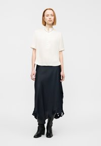 Ελαφρώς μπεζ πουκάμισο με στρογγυλό γιακά και πατιλέτα με κουμπιά σε συνδυασμό με μια αέρινη σκουρομπλε φούστα με βολάν στο τελείωμα. Μαύρες μπότες μέχρι το γόνατο.