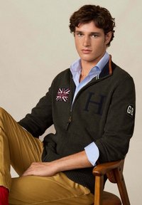 Tmavozelený svetr na zip s písmenem "H" a nášivkou Union Jack, navlečený přes světle modrou pruhovanou košili, doplněný o khaki kalhoty.