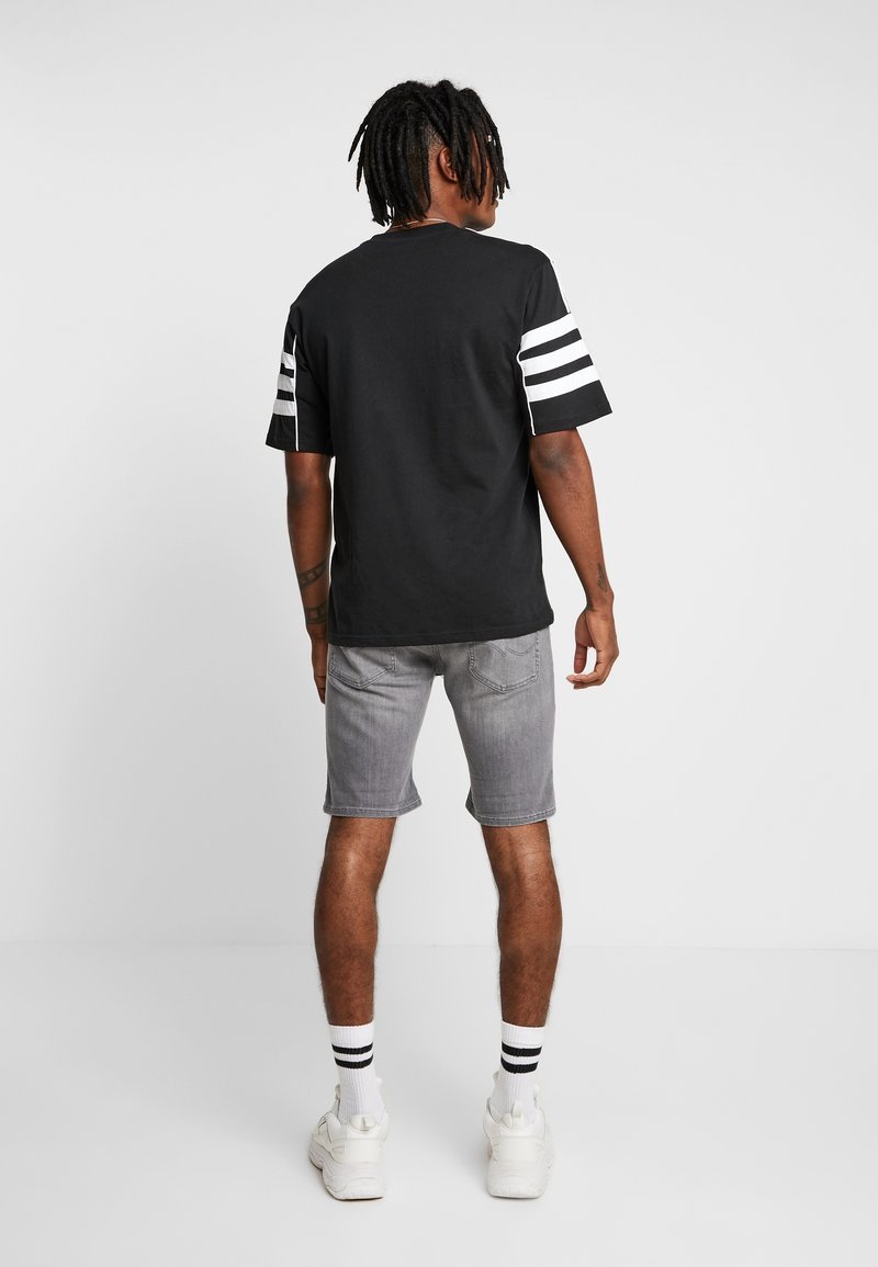 Camiseta negra con rayas blancas en las mangas, combinada con pantalones cortos grises. Modelo visto desde atrás, que lleva zapatillas blancas y calcetines a rayas.