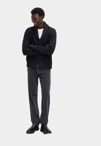 Donker gebreide cardigan met een schouderkraag, knoopsluiting, gecombineerd met grijze jeans en zwarte laarzen. Model met gekruiste armen en een lichte glimlach.