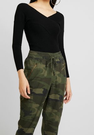 Žena v černém žebrovaném dlouhém tričku s výstřihem do V a zelených maskáčových cargo kalhotách s tkanicí v pase a kapsou na zip.