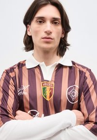 Νεαρό άτομο με καστανά μαλλιά μέχρι τους ώμους φοράει καφέ και ροζ ριγέ ράγκμπι πουκάμισο με λογότυπα, στέκεται με σταυρωμένα τα χέρια μπροστά σε απλό φόντο.