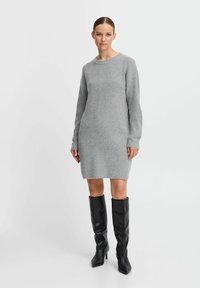 Abito in maglia grigia a coste con maniche lunghe, scollo rotondo e orlo a frange, abbinato a stivali neri sopra il ginocchio. Tessuto morbido e forma aderente.
