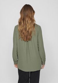 Blouse à manches longues vert olive avec une coupe décontractée, tissu doux, détail plissé au dos et poignets boutonnés, associée à un pantalon noir.