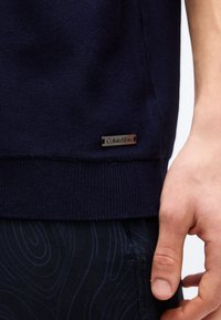 Μπλε πουαντιλέ πλεκτό τελείωμα πουλόβερ με μικρή μεταλλική ετικέτα λογότυπου Calvin Klein πάνω από σκούρο παντελόνι με σχέδια, δίπλα σε χαλαρό χέρι ατόμου.