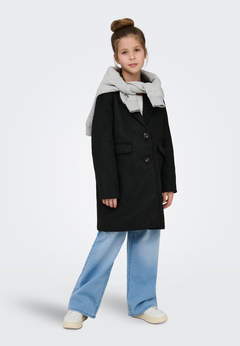 Cappotto nero con due bottoni, abbinato a una felpa grigia indossata sulle spalle. Jeans azzurri a gamba larga e sneakers bianche completano l'outfit.