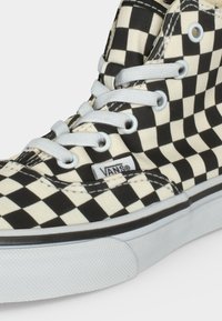 Αθλητικό παπούτσι Vans ψηλό με ασπρόμαυρο καρό σχέδιο και λευκά κορδόνια, εμφανίζει ετικέτα μάρκας στο πλάι κοντά στη λαστιχένια σόλα.