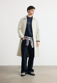 Νέος άνδρας στέκεται φορώντας μπεζ μακρύ παλτό, ναυτικό φούτερ, σκούρα φαρδιά τζιν, γκρι πουλόβερ δεμένο στη μέση και γκρι παπούτσια σε γκρι πάτωμα.