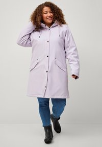 Cappotto impermeabile lilla con cappuccio, bottoni a pressione, due tasche frontali e taglio dritto, abbinato a jeans blu e stivali neri.