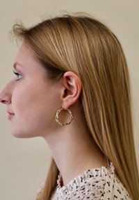 Gouden oorringen met een gedraaide, gestructureerde ontwerp, in een ronde vorm met een veilige sluiting, gedragen door een persoon met lang haar.