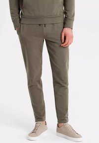 Pantalones de chándal verde olivo hechos de algodón suave, con una cintura elástica con cordón, bolsillos laterales y piernas entalladas, combinados con zapatillas beige.