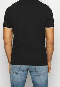 Czarna koszulka polo z krótkim rękawem, z szarym wykończeniem na kołnierzyku i rękawach, noszona z jasnoniebieskimi dżinsami. Minimalistyczny design, klasyczny krój.