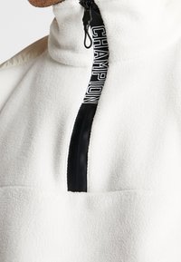 Biała polarowa kurtka z wysokim kołnierzem i czarnym zamkiem błyskawicznym oznakowana napisem "CHAMPION" w białym kolorze.