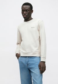 Μπεζ φούτερ με στρογγυλή λαιμόκοψη, μανίκια με μανσέτες και κεντημένο λογότυπο στο στήθος, συνδυασμένο με ανοιχτόχρωμα μπλε τζιν. Λείο ύφασμα.