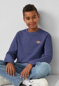 Blaues Sweatshirt mit langen Ärmeln, geripptem Kragen und orangefarbenem Diamantlogo. Getragen mit hellblauen Jeans und weißen Sneakers. Sitzende Pose.