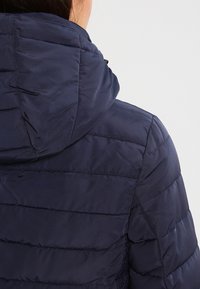 Osoba w granatowej pikowanej kurtce z kapturem, widziana od tyłu na jednolitym jasnym tle.