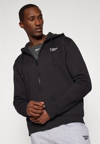 Črna zip-up kapuca s prilegajočim dizajnom, ki vsebuje logotip Reebok, kapuco na vrvico in rebraste manšete. Nosi se preko sive majice in trenirk.