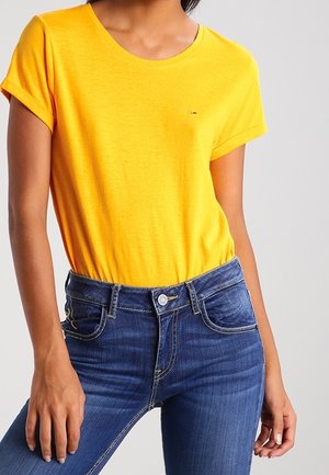 Kobieta ubrana w jasny żółty krótki rękaw T-shirt włożony w ciemnoniebieskie, wysokie spodnie jeansowe na jednolitym tle.
