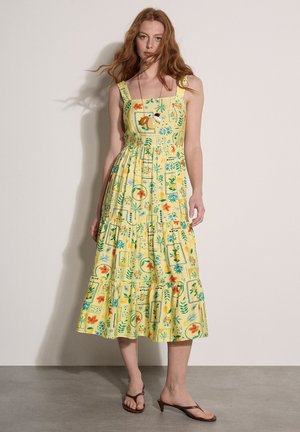 Woman standing wearing yellow sleeveless midi dress with floral and fruit print, paired with brown heeled sandals and fruit necklace.