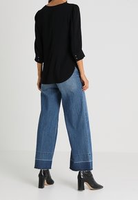 Blusa nera con maniche a tre quarti, abbinata a pantaloni in denim blu larghi con orli sfrangiati e cuciture a contrasto. Stivali neri con tacco.