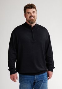 Schwarzer Zip-Pullover aus weichem Material mit langen Ärmeln und gerippten Bündchen, kombiniert mit blauen Jeans. Glatte Textur und lockere Passform.