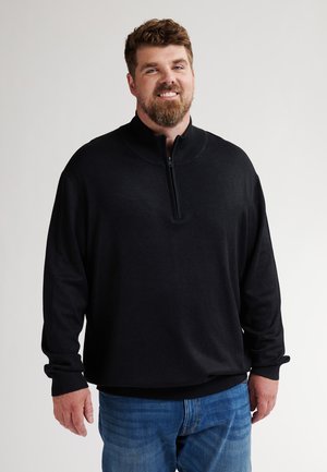 Sudadera negra con cremallera en el cuello, fabricada en un tejido suave, de manga larga y puños acanalados, combinada con unos vaqueros azules. Textura lisa y corte relajado.