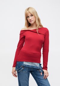 Červený top s dlouhým rukávem a odhalenými rameny, s volně splývajícím výstřihem, úzkým střihem a hladkou texturou. Kombinován s modrými denimovými džíny s unikátním designem kapes.