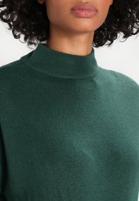 Pull à col montant de couleur vert foncé en tissu tricoté doux, avec un col côtelé et une texture lisse, vu de la poitrine vers le haut.