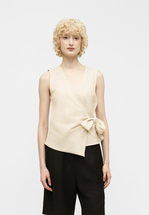 Kvinde med krøllet blondt hår iført ærmeløs beige slå-om-top med sløjfe i siden og løse sorte bukser, stående mod en ensfarvet baggrund.