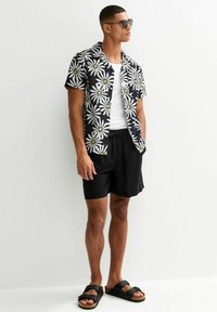 Chemise noire à manches courtes avec un motif de marguerites blanches et jaunes, assortie à un short noir et des sandales noires, offrant une coupe décontractée.