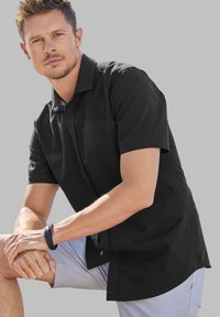 Chemise noire à manches courtes avec col, coupe décontractée et texture lisse, associée à un short de couleur claire.
